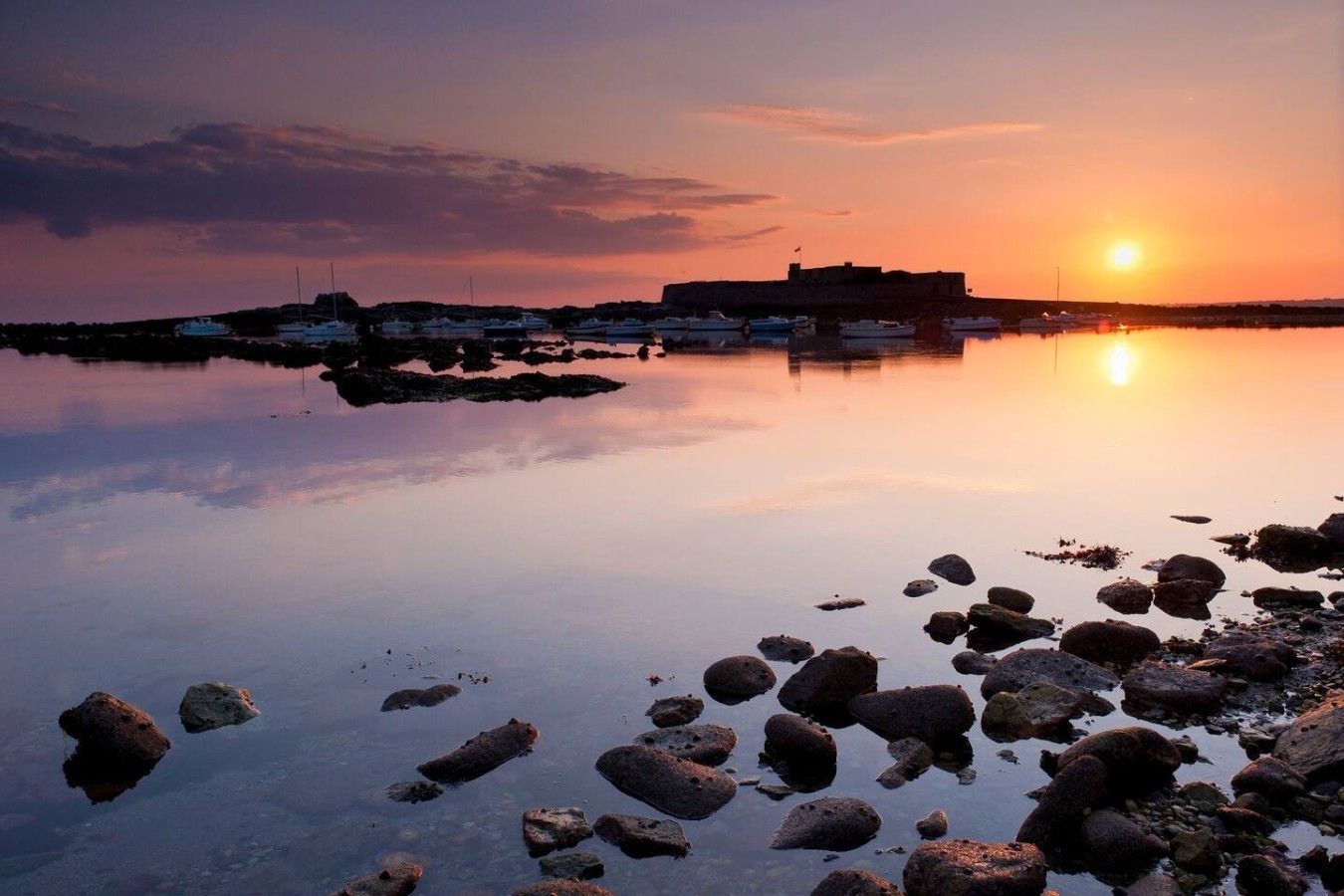 A calm waterfront with boats, a rocky shoreline, and a distant building.