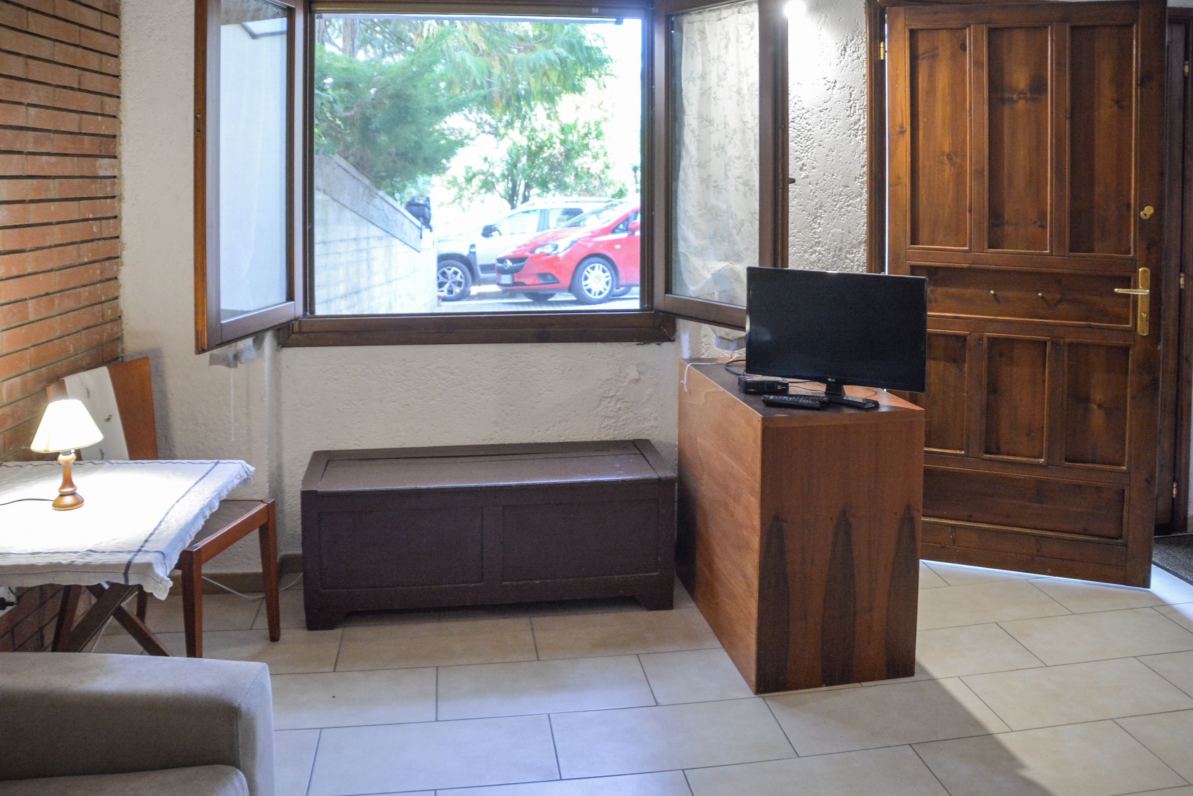 A room with a wooden desk, a television, a bench, a lamp, and a window with a view of a parked car outside.