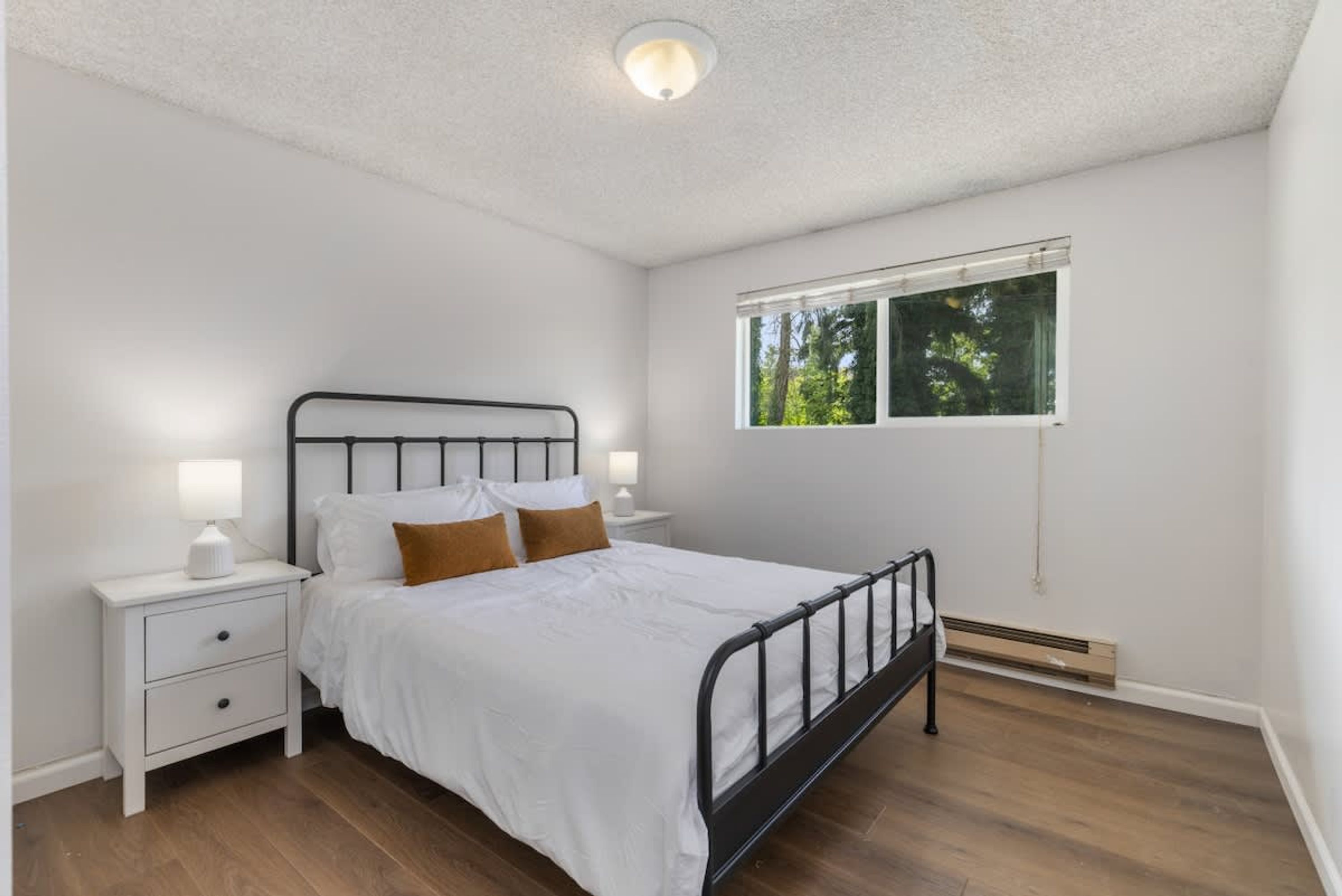 A bedroom with a single bed, two bedside tables with lamps, a window with a view of trees, and a ceiling light.