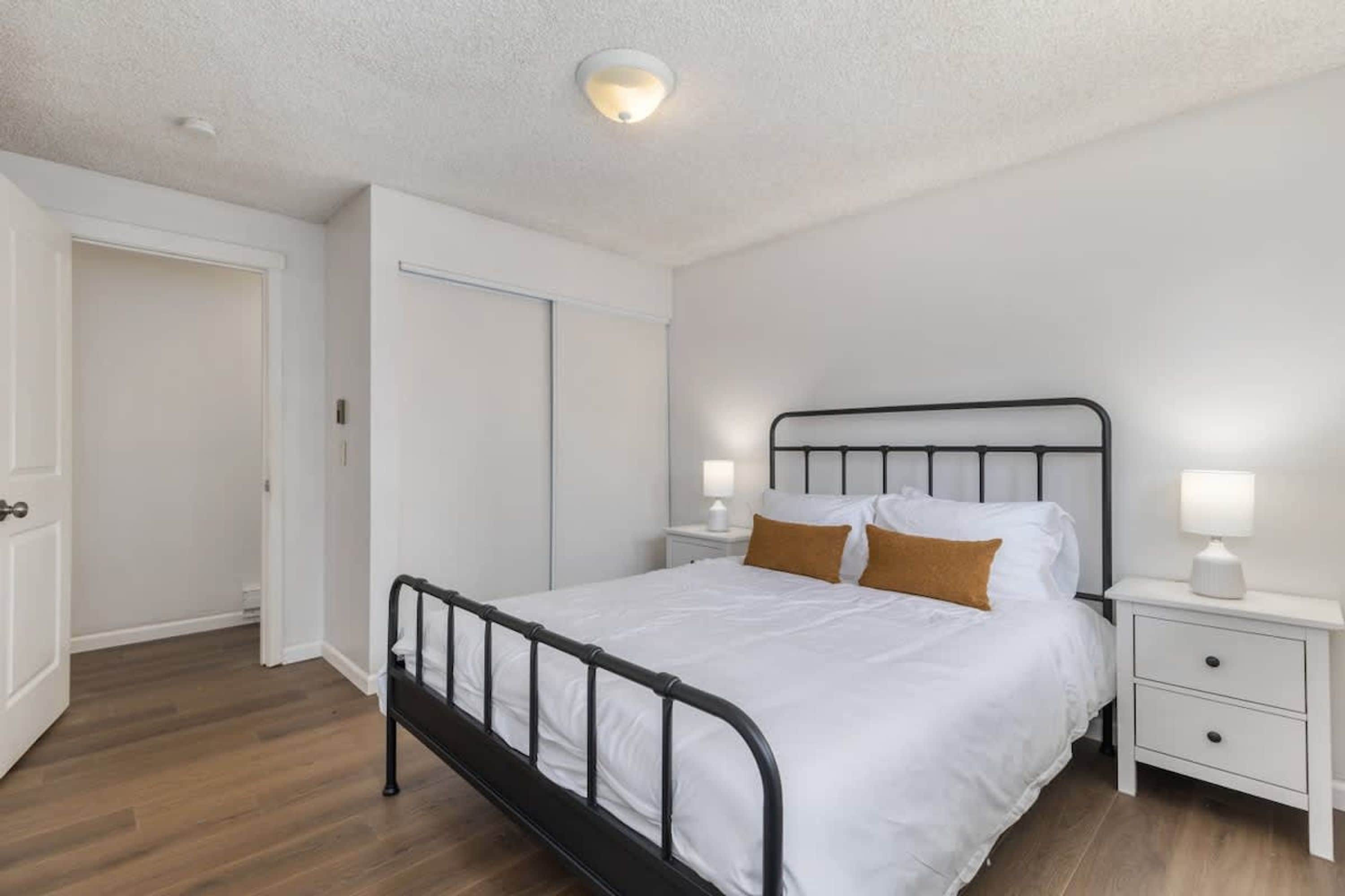 A bedroom with a single bed, white walls, a wooden floor, a white door, and a nightstand with a lamp.