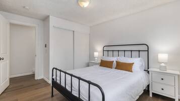A bedroom with a single bed, white walls, a wooden floor, a white door, and a nightstand with a lamp.