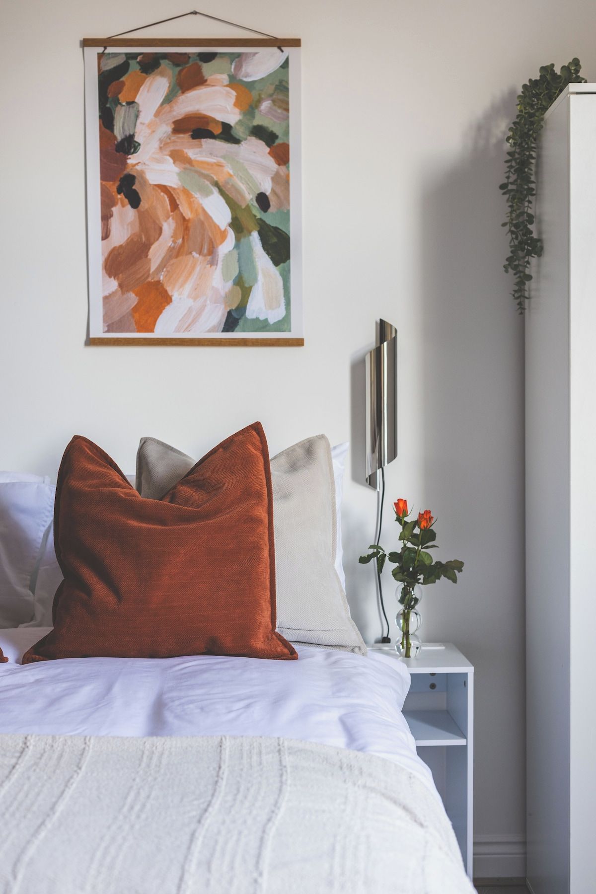 A bedroom with a bed, pillows, a bedside table with a vase of flowers, a wall-mounted artwork, and a plant.