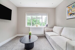 Ein modernes Wohnzimmer mit einer Couch, einem runden Couchtisch, einem Flachbildfernseher und einem Fenster mit Blick ins Grüne.