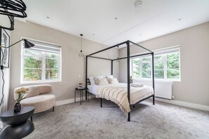 Ein Schlafzimmer mit einem Himmelbett, einer Sitzecke mit einem Sessel und einem kleinen Tisch, sowie zwei Fenstern, die für natürliches Licht sorgen.