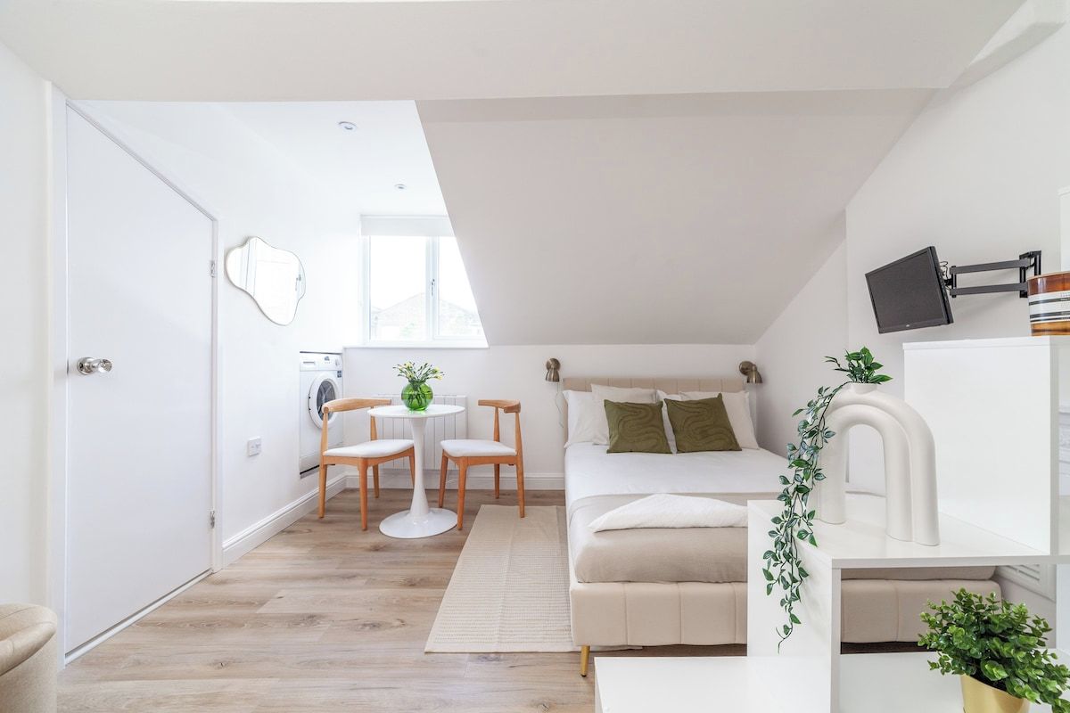 A compact living space with a bed, a small table with chairs, a washing machine, and a television mounted on the wall.