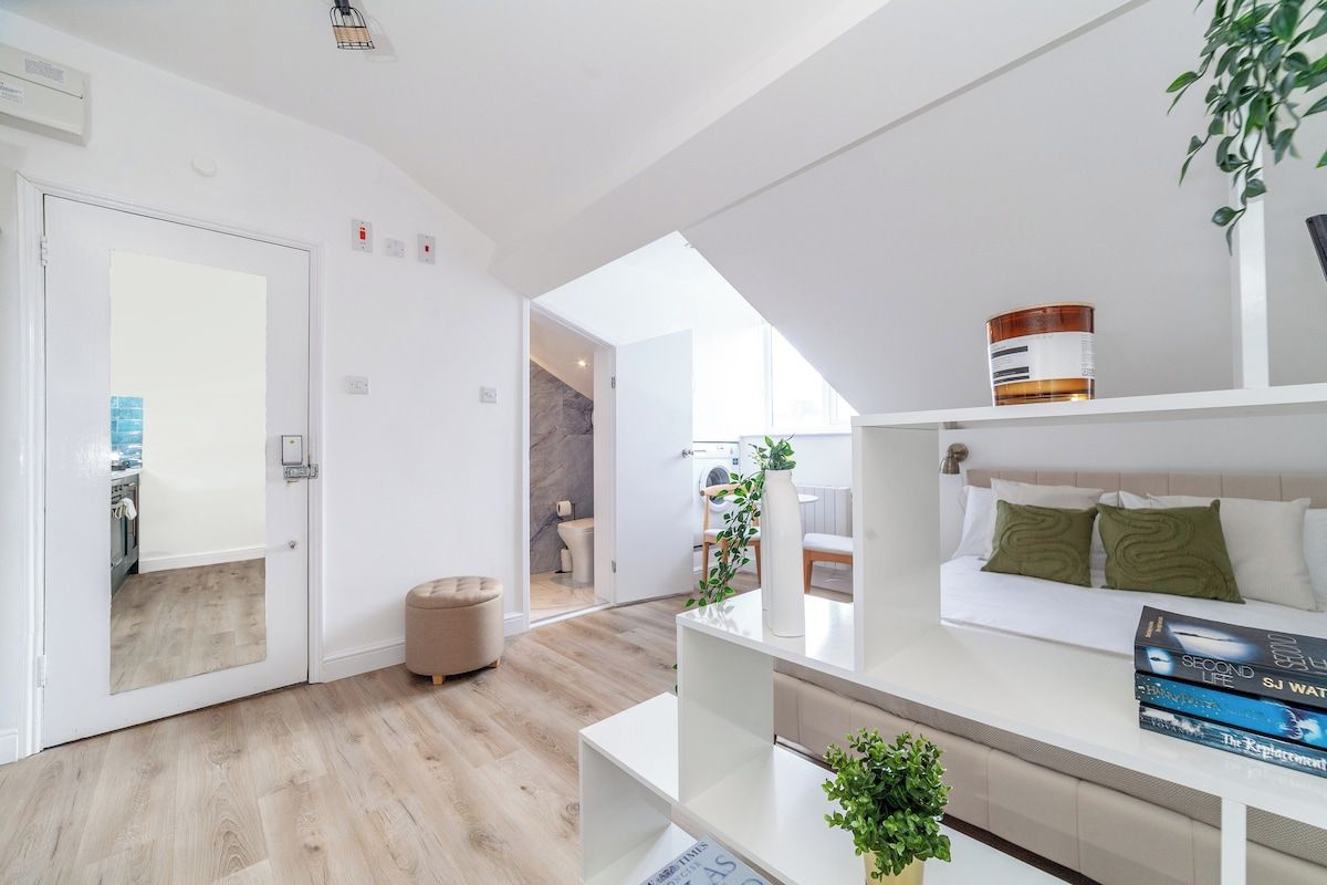 Una sala moderna con un sofá blanco, una mesa de centro y una estantería. Hay una puerta que da a una cocina y un baño.