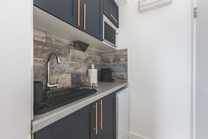 A modern kitchen with dark cabinetry, a stainless steel sink, and a built-in microwave. - Stylish cosy studio - Arsenal 15 min walk 01 (Greater London)