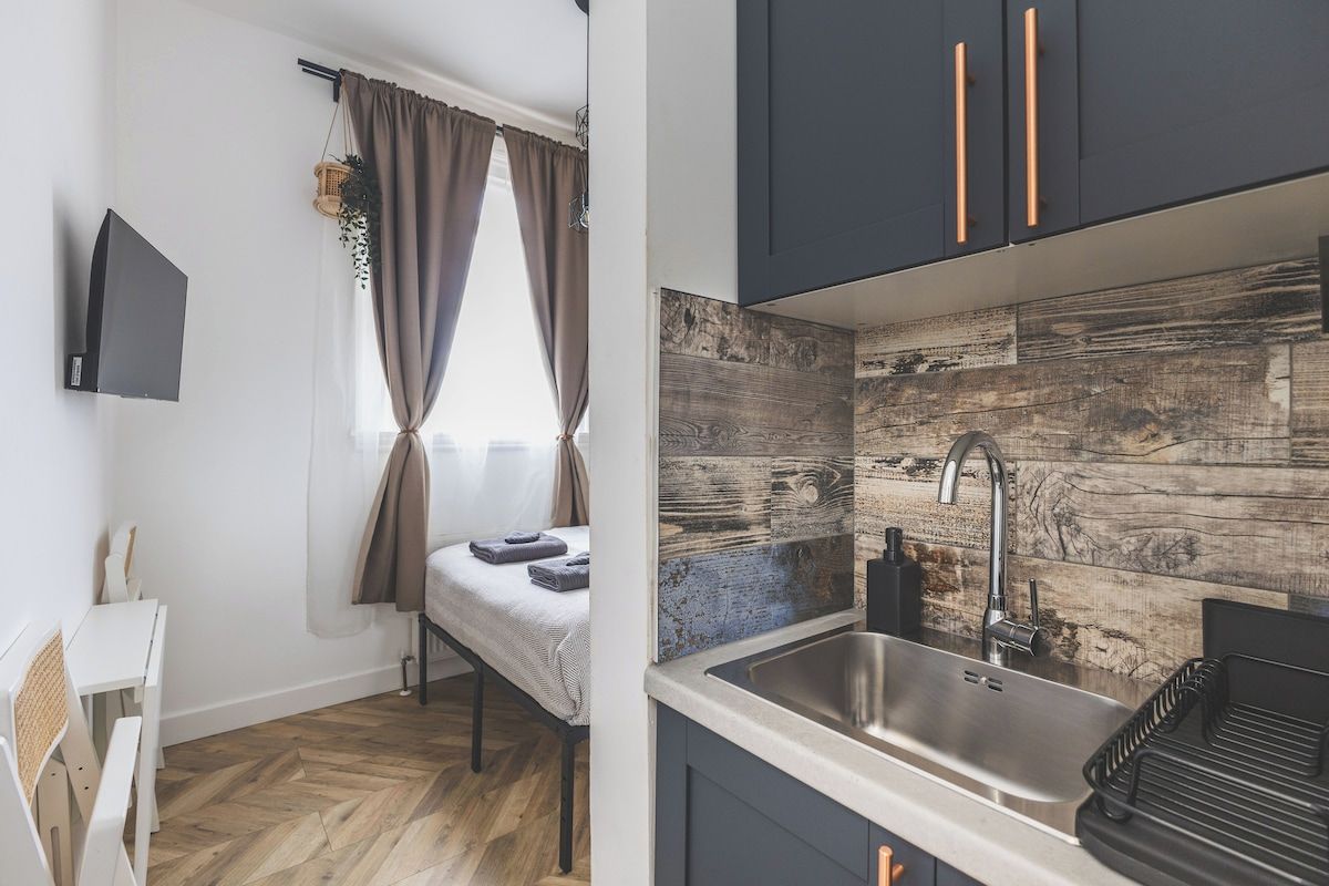 A compact kitchen with a modern design, featuring a wooden backsplash, a stainless steel sink, and dark cabinetry with copper handles.