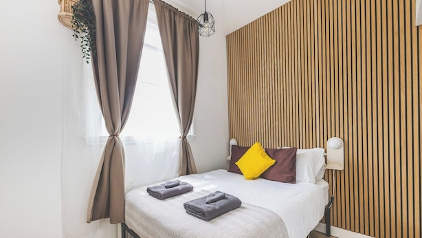 Quarto moderno com cama, cortinas, luminária na parede e painel de madeira.