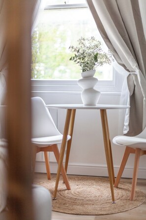 Ein Zimmer mit einem weißen Tisch und zwei Stühlen, einem Fenster mit Vorhängen und einer Vase mit Blumen auf dem Tisch.