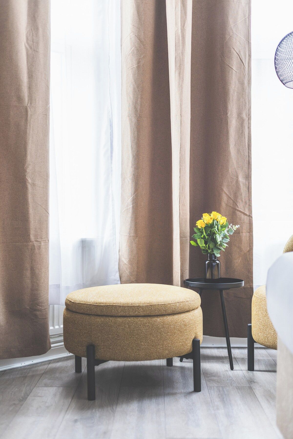 A room with a large window, a brown curtain, a round ottoman, and a small table with a vase of yellow flowers.