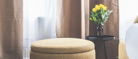 A room with a large window, a brown curtain, a round ottoman, and a small table with a vase of yellow flowers.
