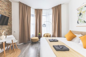 A hotel room with a bed, a desk, a chair, a small table, a lamp, and a painting on the wall. - Lovely Studio Flat in Kilburn w/great location (Greater London)