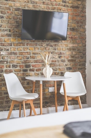 A room with a brick wall, a flat-screen TV mounted above a round table with two white chairs, and a vase with dried plants on the table. - Lovely Studio Flat in Kilburn w/great location (Greater London)