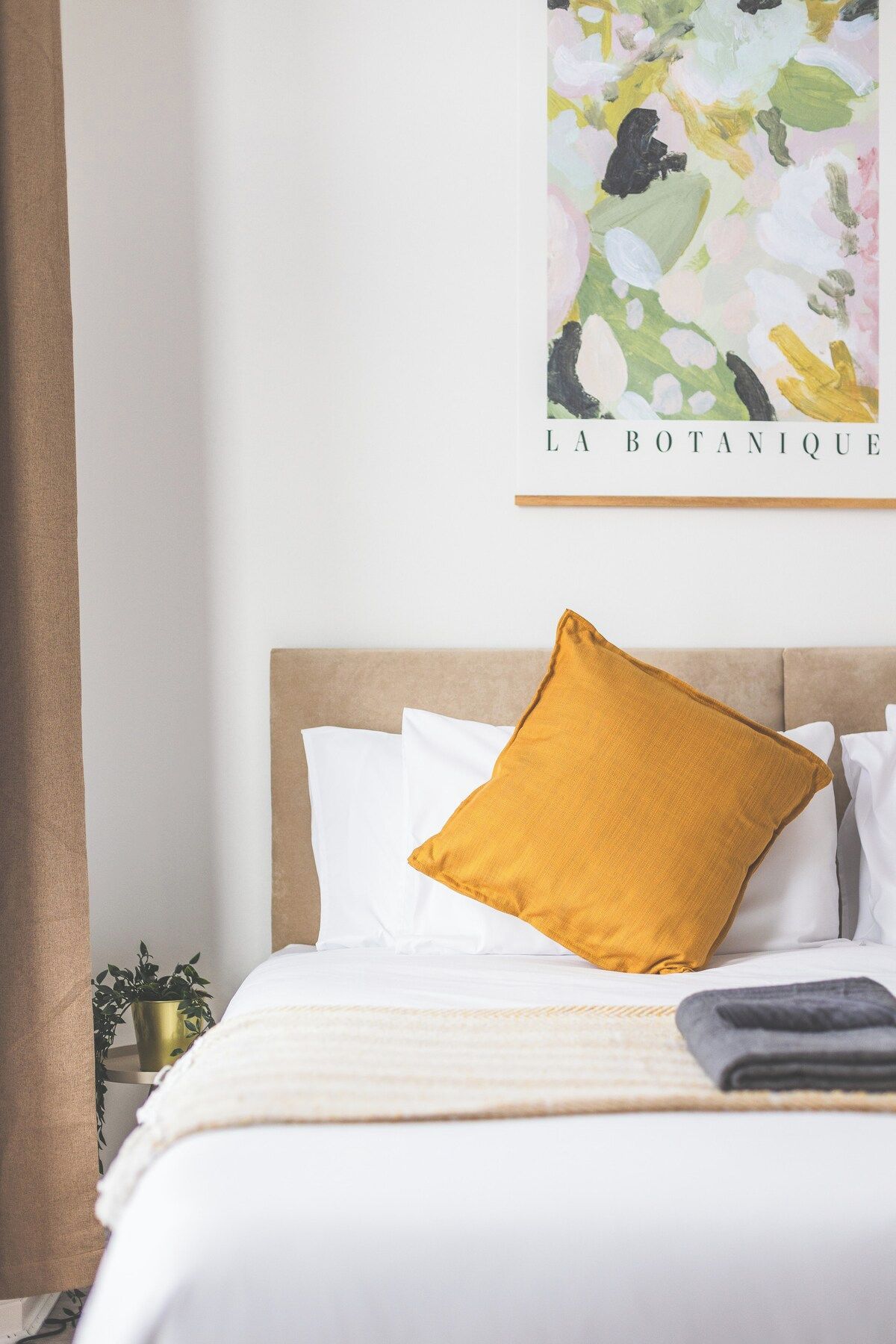 Una cama bien hecha con ropa blanca y un cojín color mostaza, una pequeña planta en maceta y una pared con una obra de arte de temática botánica.