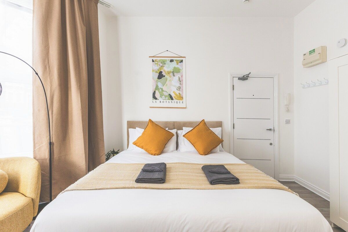Ein Hotelzimmer mit einem Bett, zwei Kissen, einem Nachttisch, einem Stuhl, einem Kleiderschrank und einem an der Wand hängenden Kunstwerk.