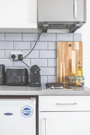 A kitchen with a white tiled backsplash, a wooden cutting board, a black toaster, a coffee maker, and a jar of olive oil. - Lovely Studio Flat in Kilburn w/great location (Greater London)