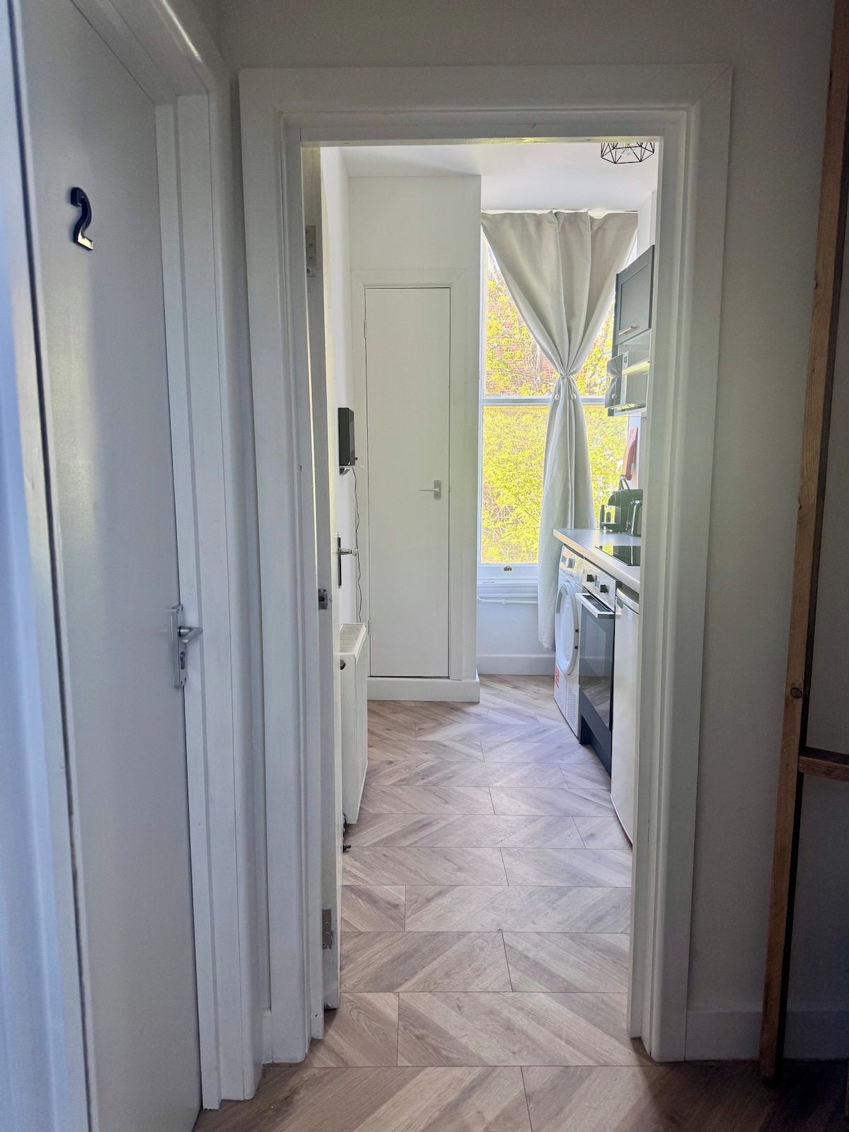 A hallway leading to a kitchen with a herringbone floor pattern, a door with a number, and a window with curtains.