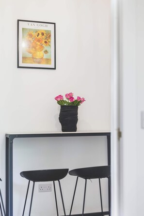 A framed artwork of sunflowers on a white wall, a black vase with pink flowers on a black table, and two black stools. - Large Studio Flat - Highbury Great Location (Greater London)