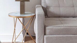 Sala de estar minimalista com sofá cinza, uma mesinha redonda com abajur e parede com discreta estampa floral.