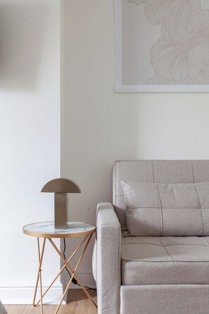 A minimalist living room with a grey sofa, a small round table with a lamp, and a wall with a subtle floral pattern. - Chic & Comfy Studio for 3 Guests, Finsbury Park (Greater London)