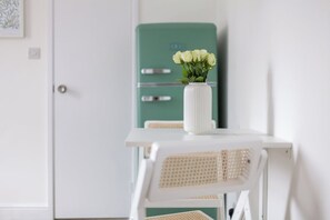 A white table with a white vase containing white flowers, a green vintage-style refrigerator, and a white chair with a woven backrest. - Gorgeous Studio Flat, Finsbury Park (Greater London)