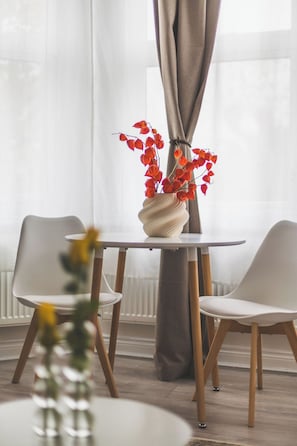 A dining area with a round table, two chairs, a vase with red flowers, and a window with sheer curtains. - Fabulous Studio Flat Kilburn - Central Location (Greater London)