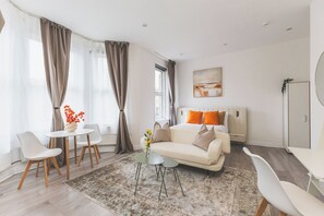 A modern hotel room with a bed, a small table, two chairs, a sofa, and a large window with curtains. - Fabulous Studio Flat Kilburn - Central Location (Greater London)