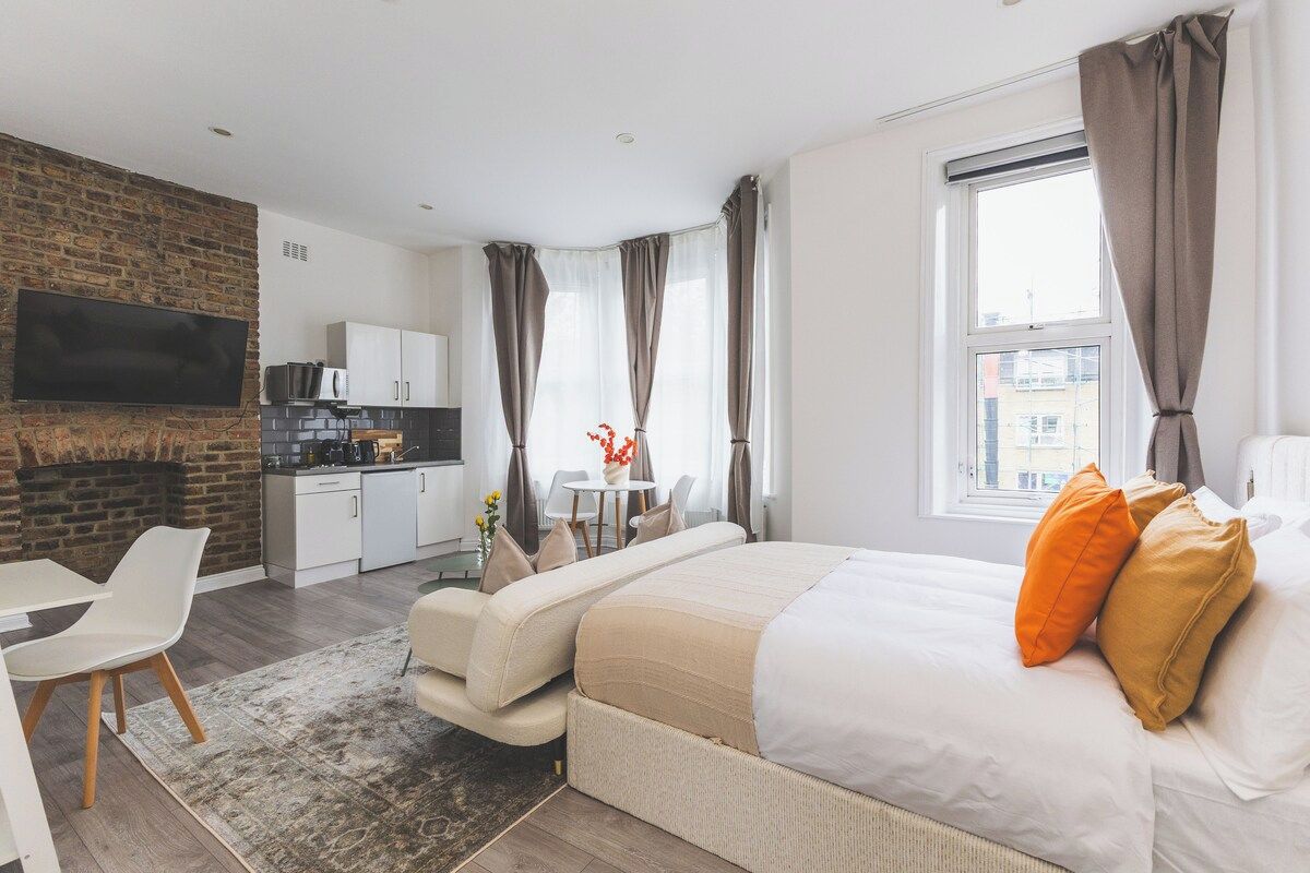 Et moderne hotelværelse med et fladskærms TV monteret på en murstensvæg, et tekøkken, en sofa, en seng med orange puder og et stort vindue med gardiner.