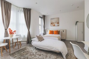 A modern bedroom with a bed, bedside tables, a chair, and a large window with curtains. - Fabulous Studio Flat Kilburn - Central Location (Greater London)