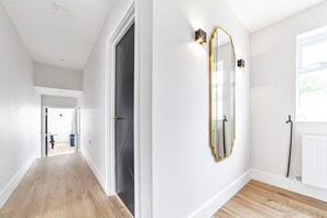 Een gang met een houten vloer, een deur naar een andere kamer, en een spiegel met gouden lijst aan de muur.