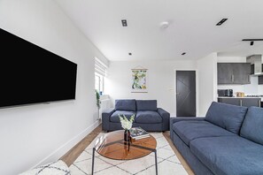 Een moderne woonkamer met een flatscreen televisie, donkere banken, een salontafel en een keuken in de achtergrond.