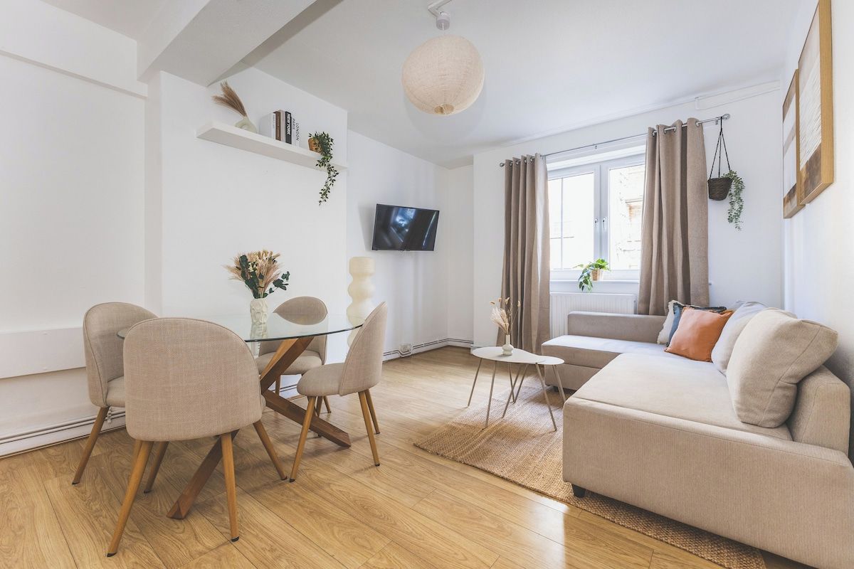 A modern living room with a glass dining table, beige sofas, and a television mounted on the wall.