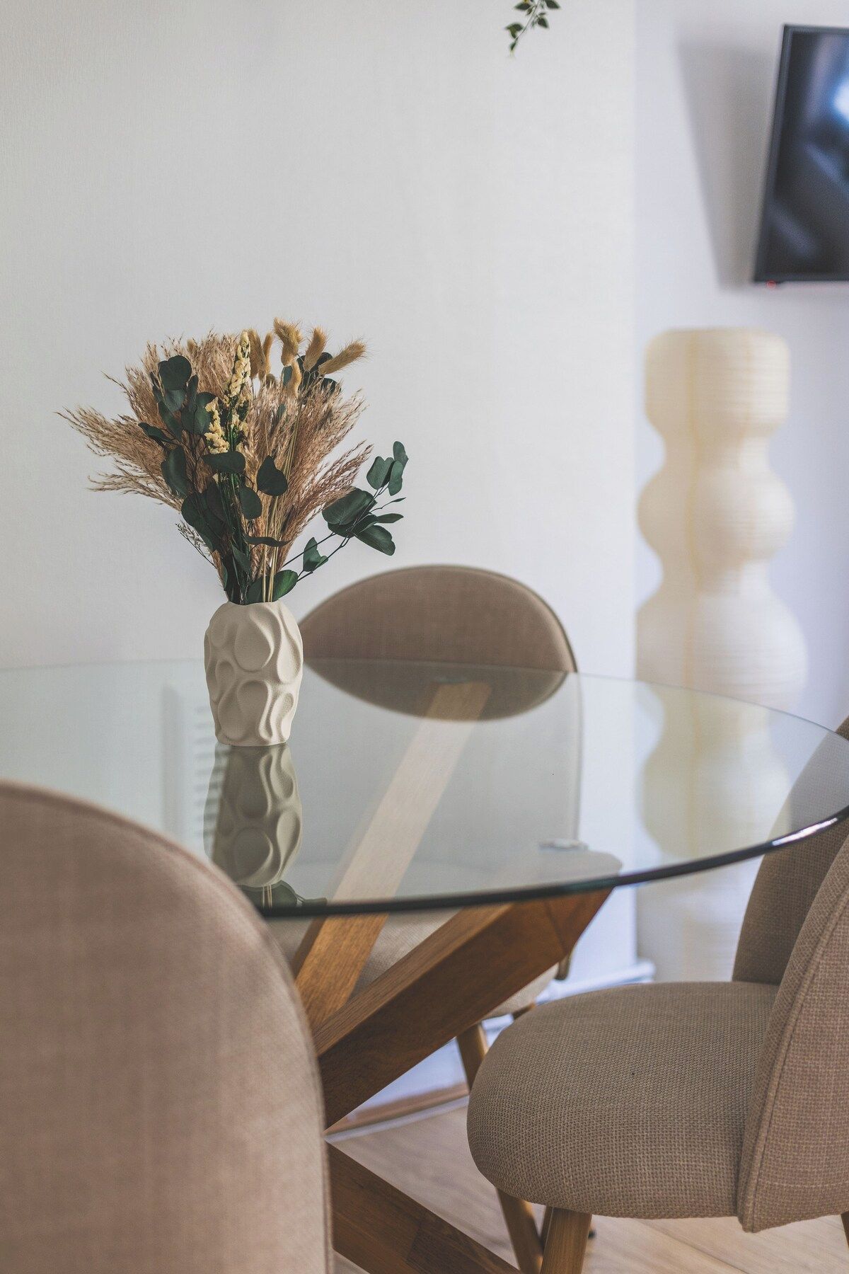 A modern dining area with a glass table, two chairs, and a vase with dried plants.
