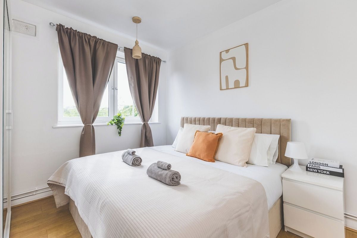 A hotel room with a bed, a nightstand, a window with curtains, and a wall-mounted artwork.