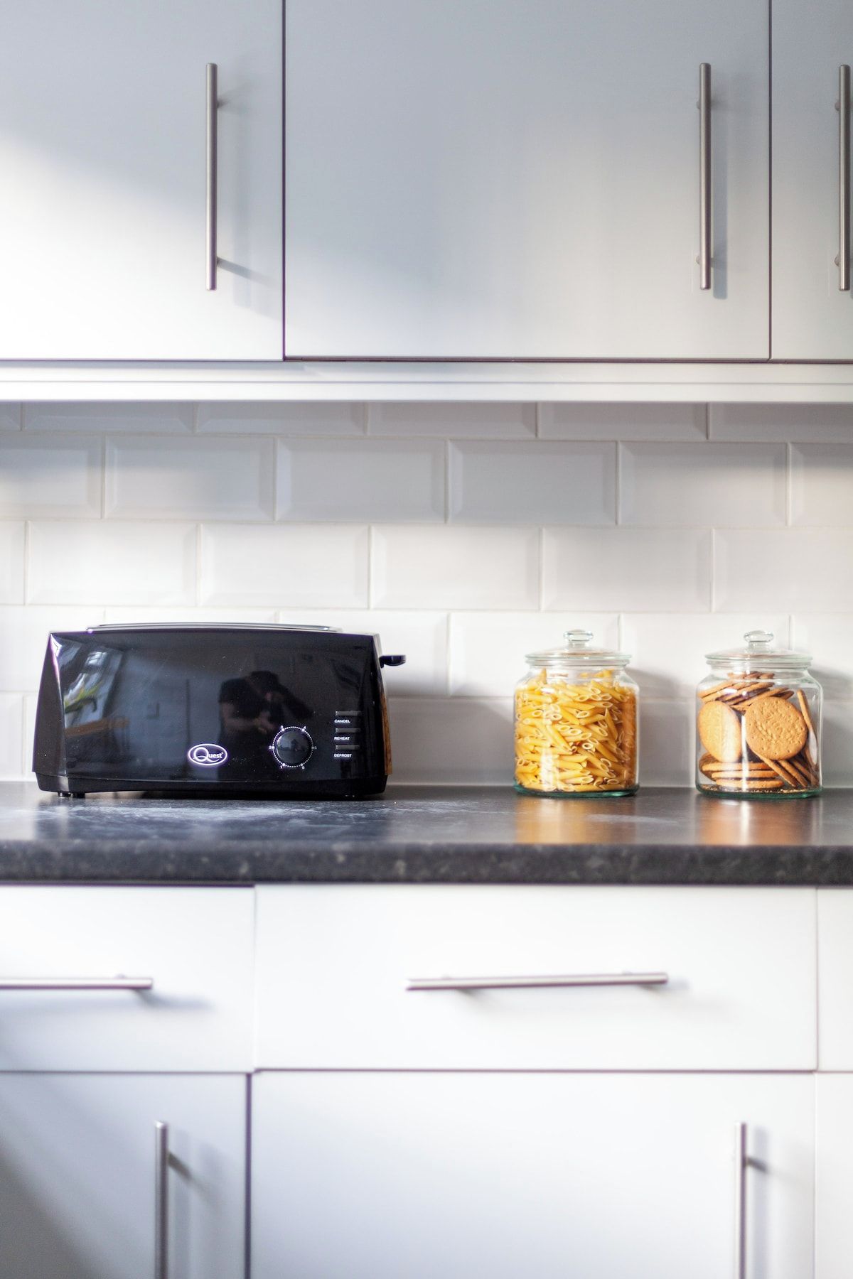 A kitchen with a toaster, two glass jars with pasta and cookies, and white cabinets.