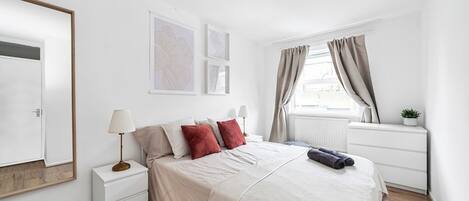 Une chambre à coucher avec un lit, des tables de chevet, une fenêtre avec des rideaux et des œuvres d’art murales.