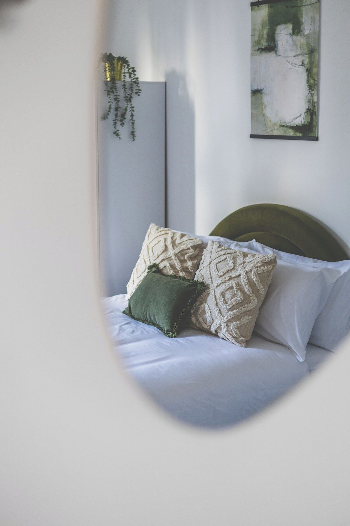 A bedroom with a bed, pillows, a headboard, a wall-mounted artwork, and a hanging plant.