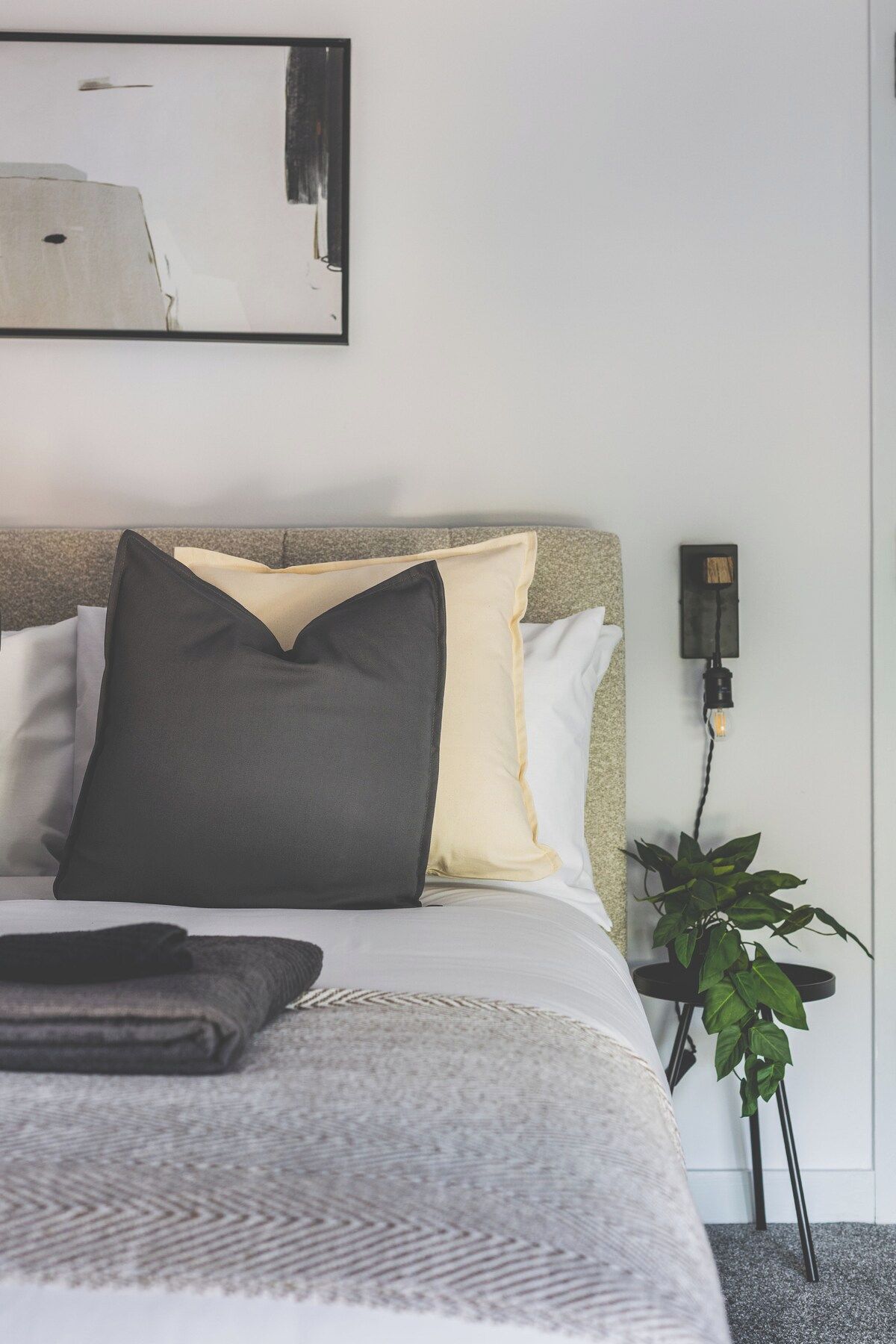 A neatly made bed with a grey and white duvet, a black pillow, and a white pillow. There is a small bedside table with a plant and a wall-mounted light.