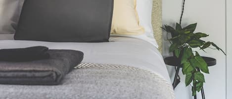 A neatly made bed with a grey and white duvet, a black pillow, and a white pillow. There is a small bedside table with a plant and a wall-mounted light.
