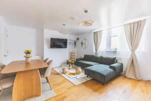 A modern living room with a wooden dining table, a green sofa, and a flat-screen TV mounted on the wall. - New Spacious 3 bed flat Duplex, Whitechapel (Greater London)