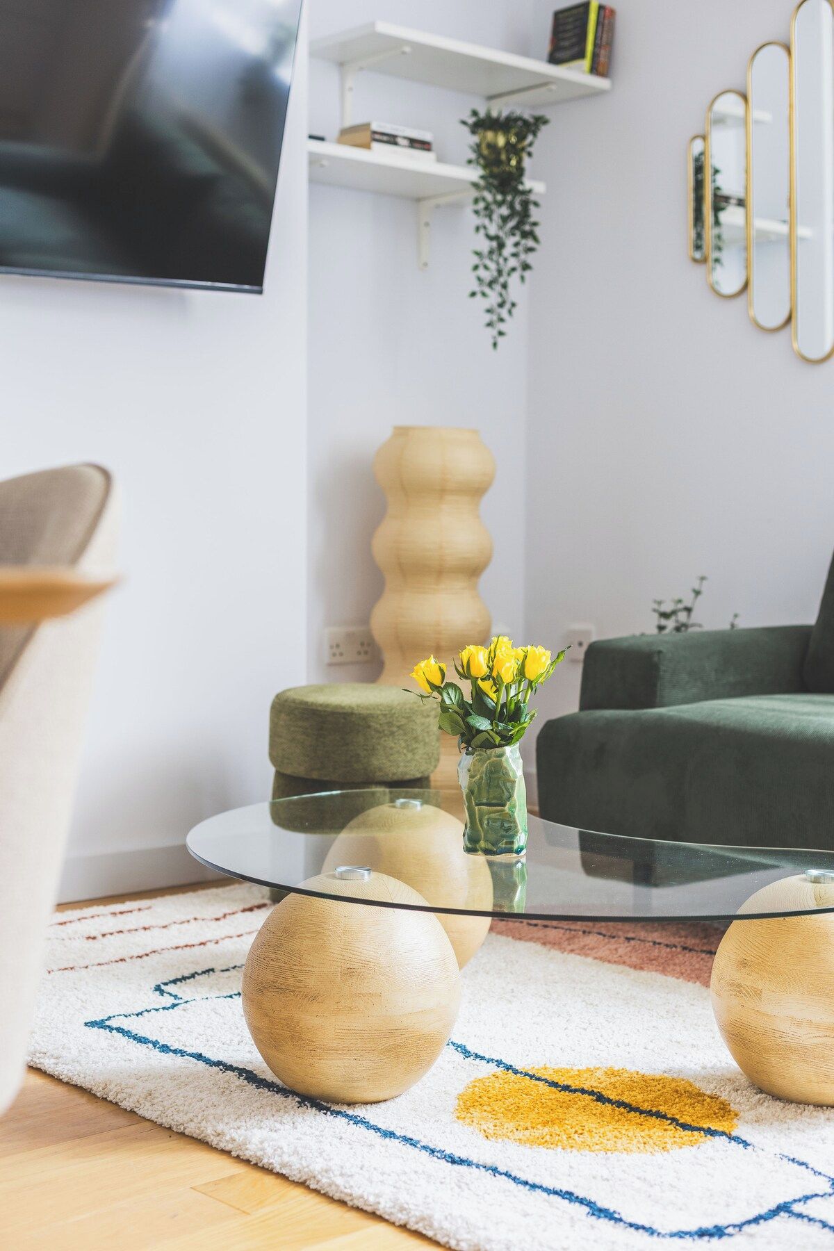 A modern living room with a glass coffee table, a green sofa, and a wooden side table with a vase of yellow flowers.