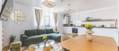 A modern living space with a sofa, coffee table, and dining table, kitchen area with cabinets and appliances, and a large window with curtains.