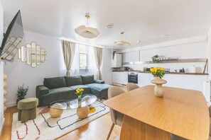 A modern living space with a sofa, coffee table, and dining table, kitchen area with cabinets and appliances, and a large window with curtains. - New Spacious 3 bed flat Duplex, Whitechapel (Greater London)