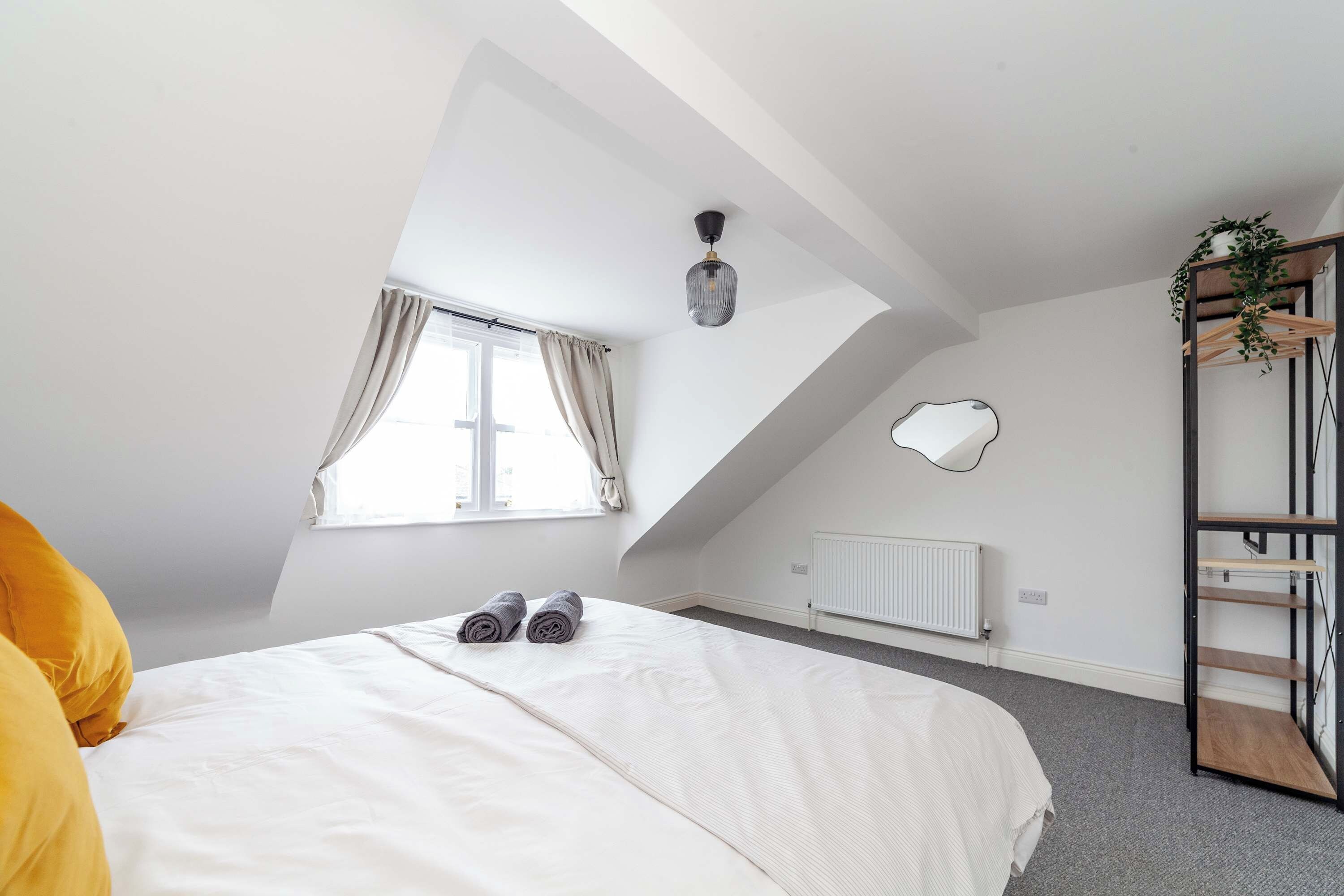 A bedroom with a sloped ceiling, a bed with rolled towels, a radiator, a hanging plant, and a wardrobe with shelves.