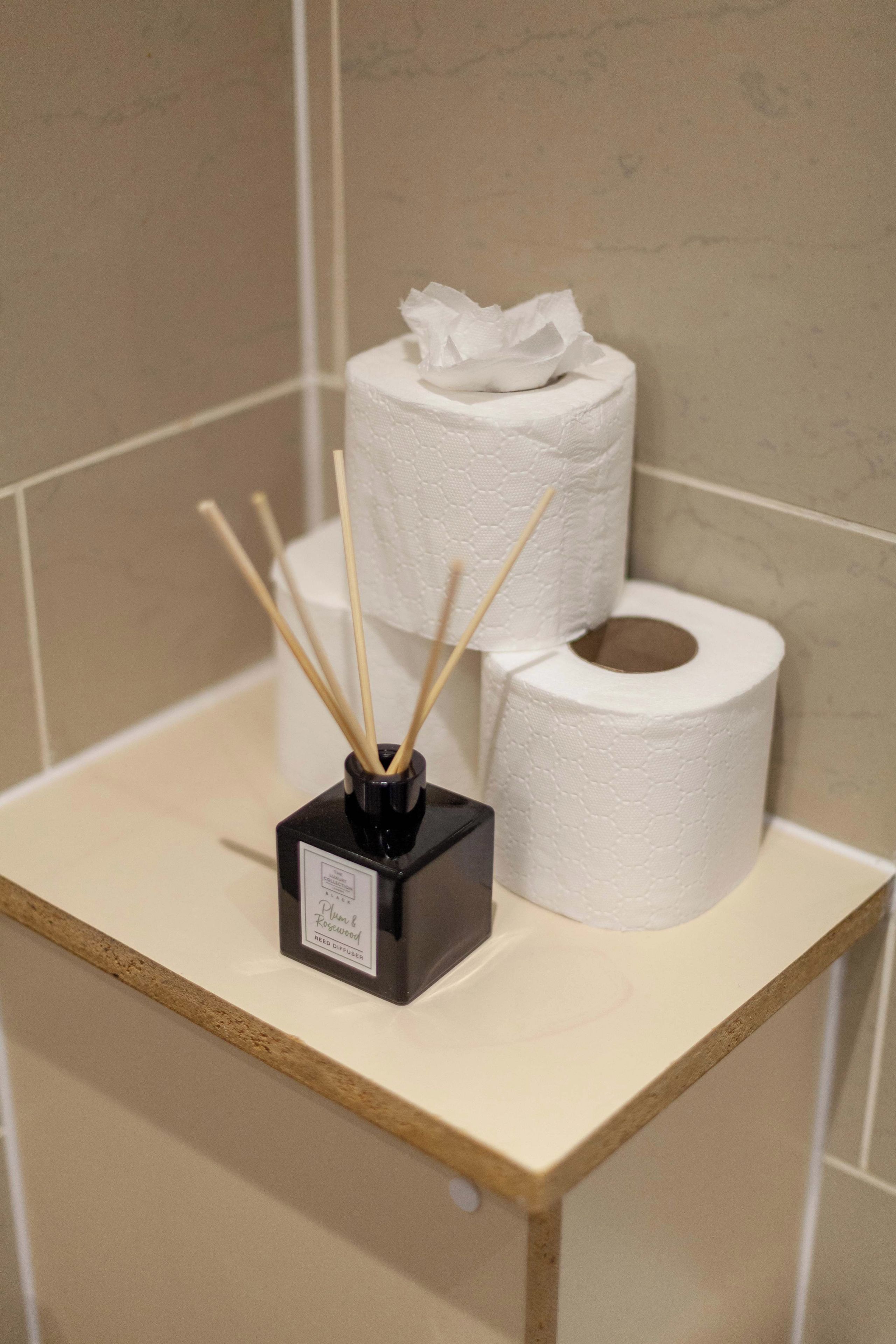 Un estante de baño con un difusor de caña, dos rollos de papel higiénico y una caja de pañuelos.