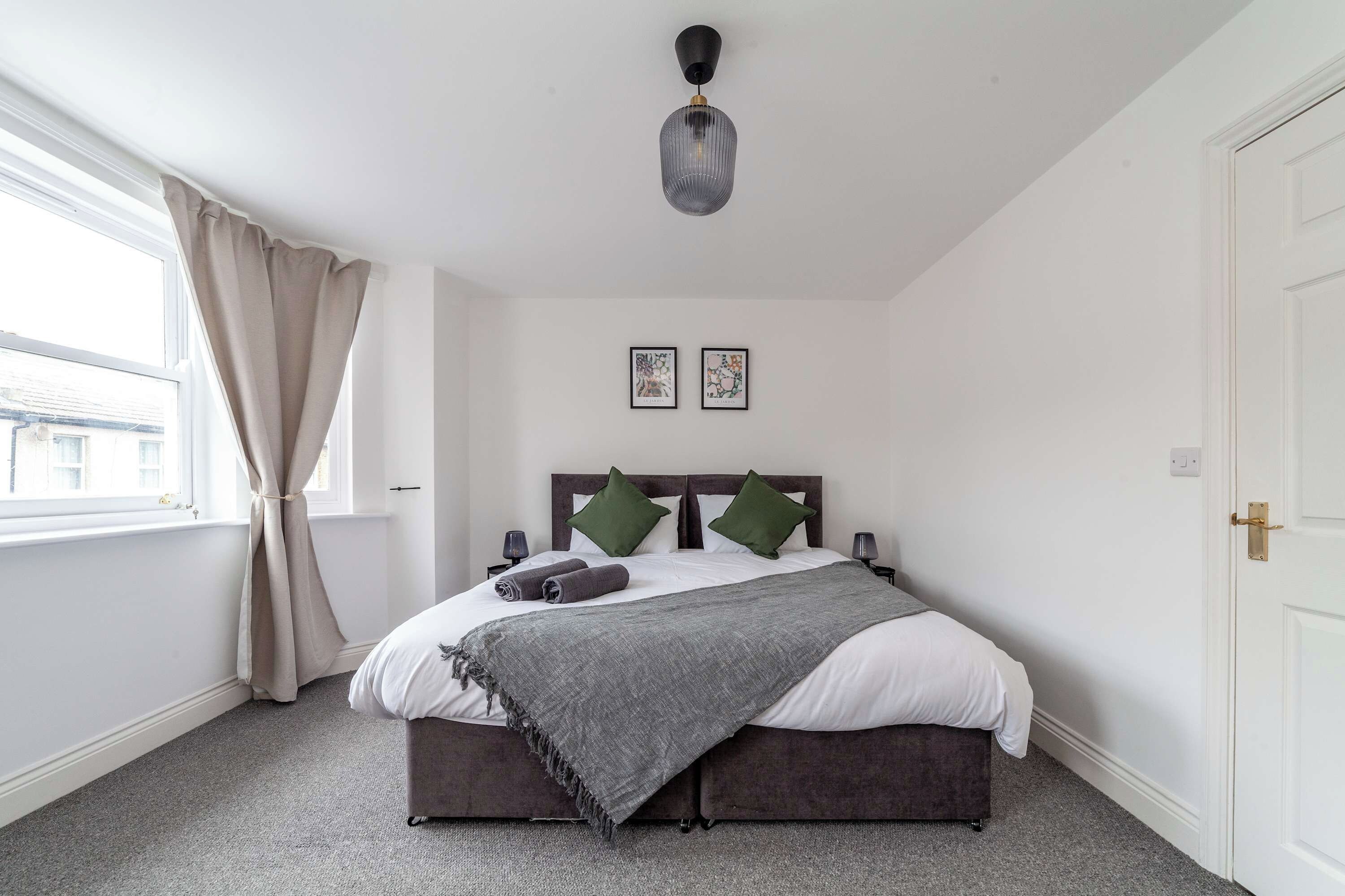Een slaapkamer met een bed, een raam met gordijnen, een wandlamp en twee ingelijste foto's.