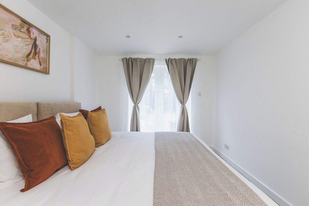A hotel room with a bed, pillows, a painting on the wall, and a window with curtains.