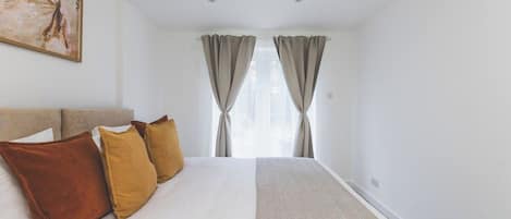 A hotel room with a bed, pillows, a painting on the wall, and a window with curtains.