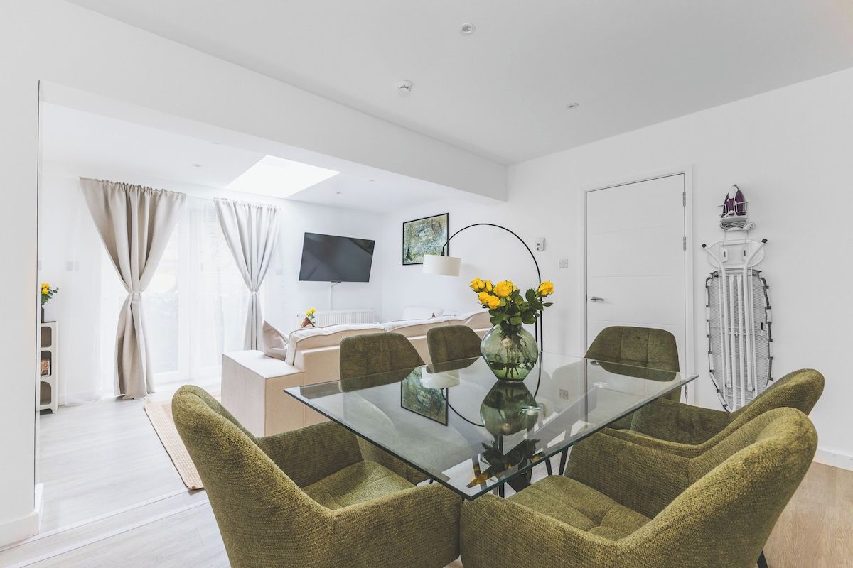 A modern dining area with a glass table and green upholstered chairs, a white sofa, and a television in the background.